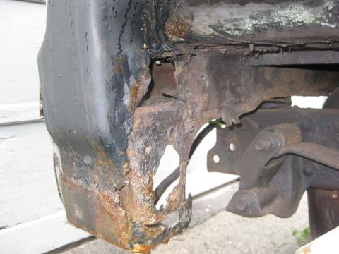 1983 Ford Bronco - Low underbody shot of a severely rusted right-side tub support and body-mount bracket, the sheet metal rotted through into jagged holes wit…