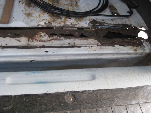 1983 Ford Bronco - Looking down into the rear body tub of a white vehicle where the inner tub support rail has rusted through, leaving a ragged row of holes w…