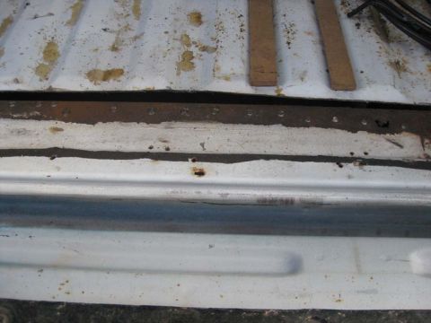 1983 Ford Bronco - Overhead close-up of a Bronco's rear tub support and rocker area, showing the ribbed white floor pan with dirt and surface rust, a rusted m…