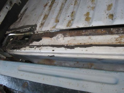1983 Ford Bronco - Close-up along the tub support rail at the edge of a white ribbed floor pan, showing heavy surface rust, flaking paint, black tar sealer an…