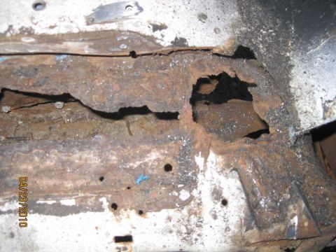1983 Ford Bronco - Extreme close-up of severe rust perforation where a tub support rail meets the white body floor, with layered scale, a large ragged hole an…