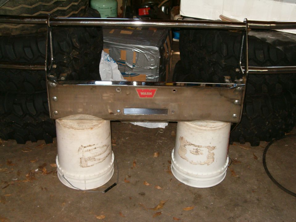 Winch bumper with WARN logo, supported by two white buckets, with tires in the background