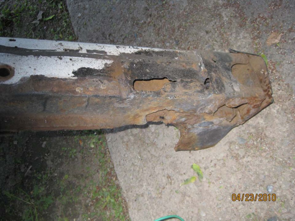 Heavily rusted metal frame part with holes and jagged edges, resting on concrete and grass