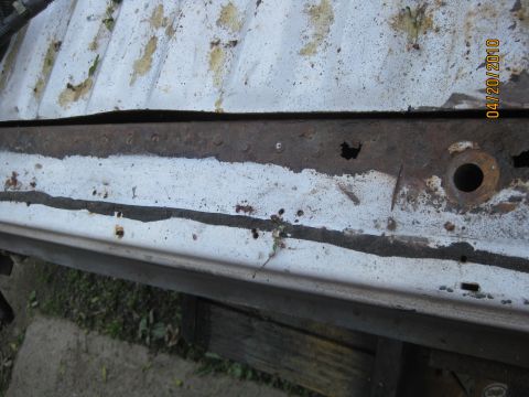 1983 Ford Bronco - Close-up along a white ribbed cargo floor and the rusty spot-welded tub support rail beneath it, showing a rust-eaten hole in the rail and…