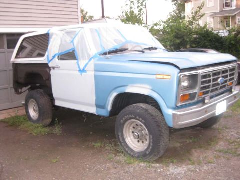 1983 Ford Bronco - Three-quarter front view of a light blue full-size Ford Bronco mid-bodywork in a gravel driveway, with the cab and glass masked off in plas…