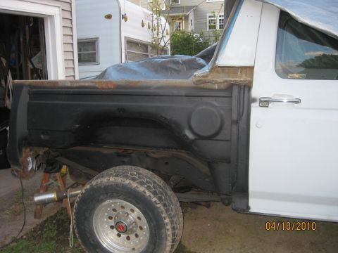 1983 Ford Bronco - Driver's-side view of a white Bronco with the entire outer rear quarter panel cut off, leaving the black inner tub wall, surface rust along…