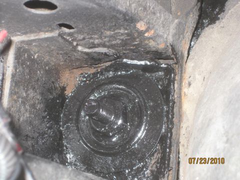 1983 Ford Bronco - Close-up of a body mount bushing and its threaded stud bolt seated in a rusty, grease-blackened frame pocket, shot from directly above with…