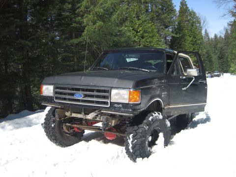 1989 Ford Bronco - A lifted black 1989 Ford Bronco navigates a snowy forest road with its driver's door ajar.