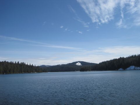1989 Ford Bronco - A wide shot of a tranquil lake surrounded by dense evergreen forests and rolling hills, with patches of snow visible on the higher elevatio…