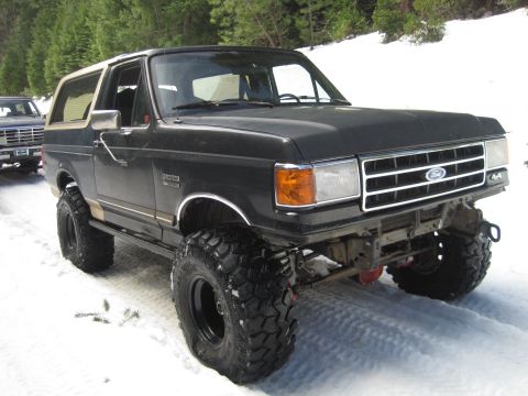 1989 Ford Bronco - A black Ford Bronco with a tan top and oversized tires sits in a snowy landscape, with another vehicle visible behind it.