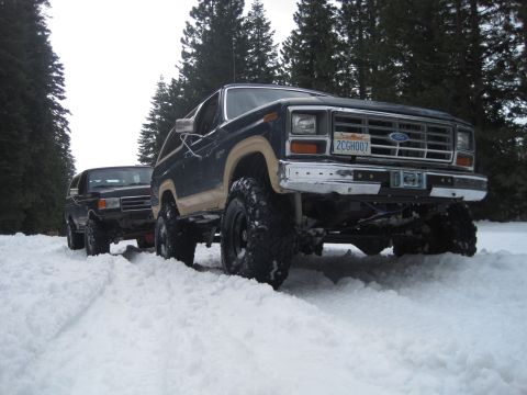 1989 Ford Bronco - Two Ford Broncos navigate a snowy trail surrounded by tall evergreen trees.