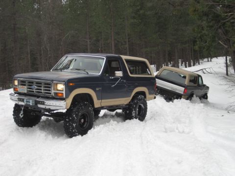 1989 Ford Bronco - Two Ford Broncos navigate a snowy trail, with one vehicle appearing to be stuck in deep snow.