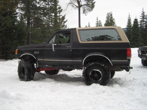 1989 Ford Bronco - Side view of a lifted 1989 Ford Bronco with oversized tires, parked in a snowy, wooded area.