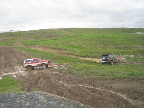 1989 Ford Bronco - Two off-road vehicles traverse a muddy terrain under an overcast sky.
