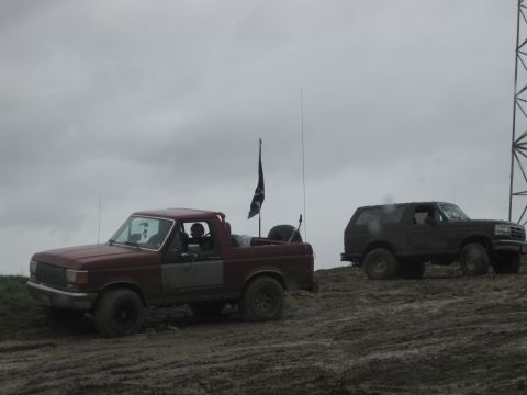 1989 Ford Bronco - Two Ford Broncos, a red one and a dark one, are parked on a muddy incline under an overcast sky.