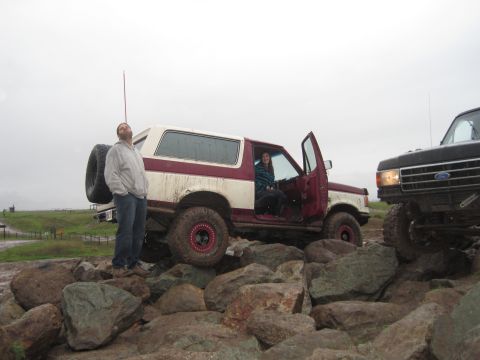 1989 Ford Bronco - A man stands next to a maroon and white Ford Bronco while another vehicle is parked nearby on a rocky terrain.