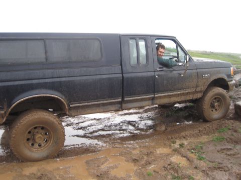 1989 Ford Bronco - A black pickup truck with a camper shell navigates a muddy terrain, showing an F250 badge on its fender.