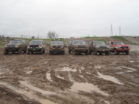 1989 Ford Bronco - A group of modified trucks and SUVs are lined up in a muddy, off-road area.