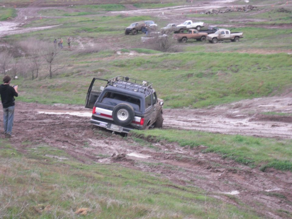 Dark 1991 Ford Bronco stuck in mud with door open, roof rack, and spare tire on rear