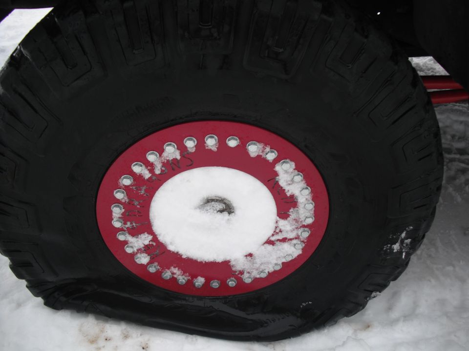 Close-up of a large off-road tire with a red beadlock ring and snow in the center. Some writing is visible on the beadlock