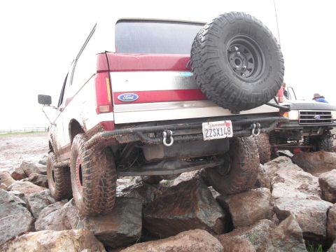 1991 Ford Bronco - A red and white Ford Bronco navigates a rocky terrain, with another Ford truck visible behind it.