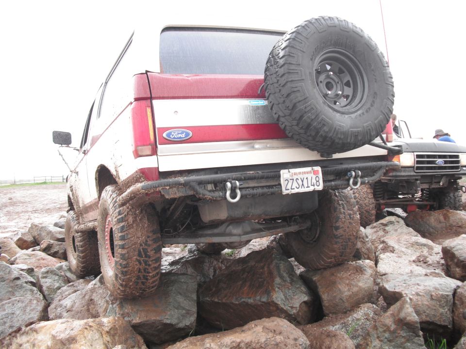 Red and white Ford Bronco with spare tire on rear, driving over rocks