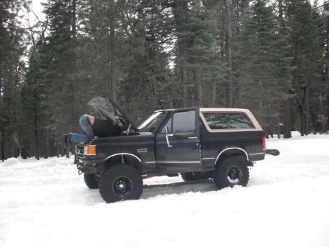 1991 Ford Bronco - A person works on the engine of a black 1991 Ford Bronco with a tan top, parked in the snow.
