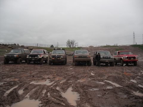 1991 Ford Bronco - A line of six Ford Broncos parked in a muddy field under an overcast sky.