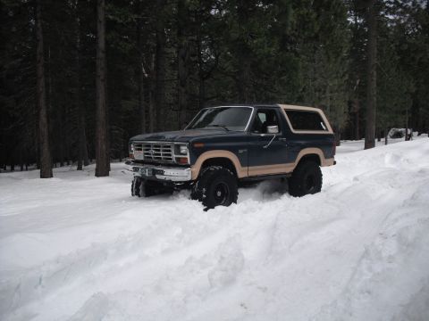 1991 Ford Bronco - A lifted Ford Bronco navigates a snowy forest road, kicking up snow as it drives.