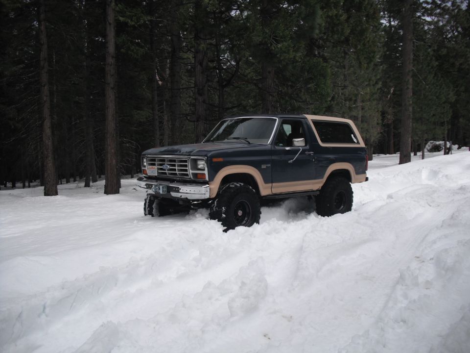 Dark blue Ford Bronco with tan trim driving through deep snow, lifted suspension, off-road tires