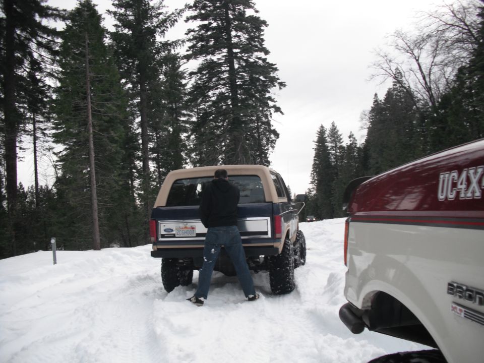 Blue Ford Bronco with tan top in snow, person standing behind it. Another white truck is to the right