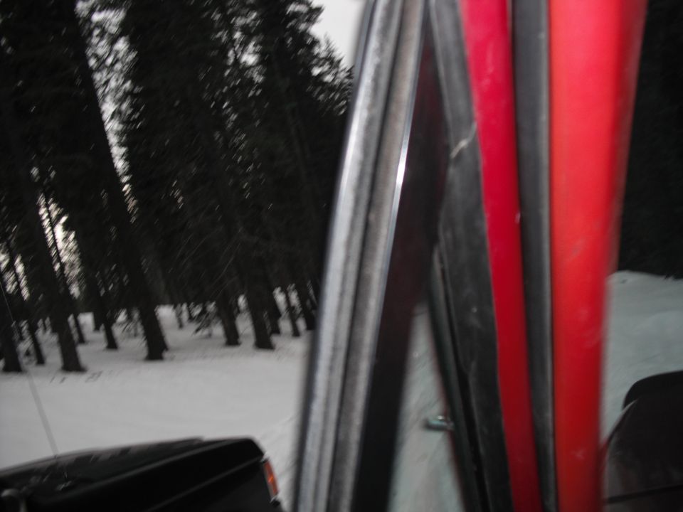 View from inside a red and black vehicle, looking out at a snowy forest. Another black vehicle is partially visible