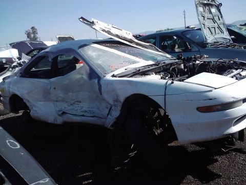 1995 Ford Probe - A severely damaged white coupe, likely a 1993 Ford Probe, sits in a junkyard with its hood open.