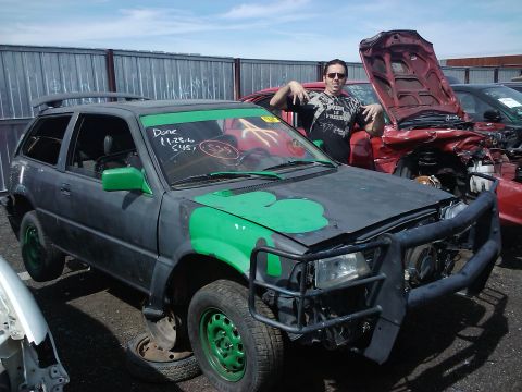 1995 Ford Probe - A heavily modified hatchback with green shamrock paint and a bull bar sits in a junkyard next to other damaged vehicles.