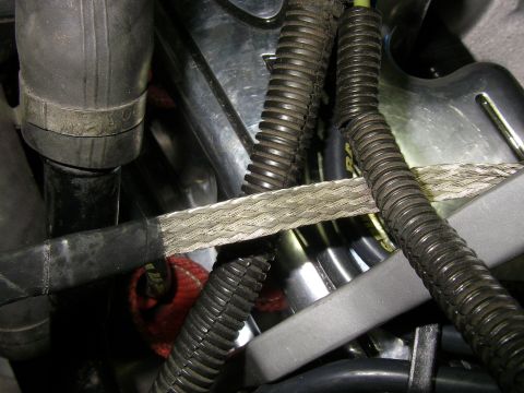 1990 Ford Bronco - Tight engine-bay shot of a wide flat braided-metal grounding strap routed across black corrugated wiring loom and rubber hoses. The strap i…