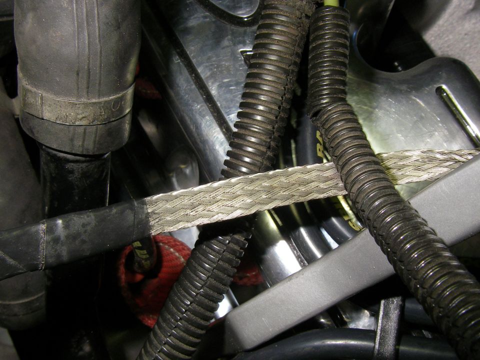 Braided flat metal grounding strap routed across corrugated wiring loom in an engine bay