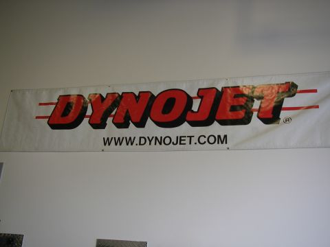 1990 Ford Bronco - White vinyl DynoJet banner with red and black block lettering and 'WWW.DYNOJET.COM' hung on an interior wall above diamond-plate trim; no v…