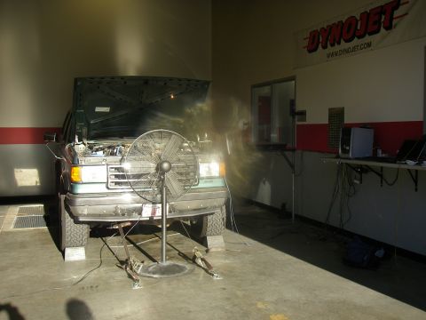 1990 Ford Bronco - A teal-green square-body Ford SUV sits strapped down on a chassis dyno with its hood open and headlights on, a large pedestal cooling fan a…