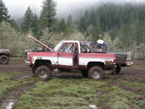 1978 Ford Bronco - Two people are working on a lifted pickup truck in a muddy, wooded area.