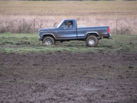 1978 Ford Bronco - A blue pickup truck navigates a muddy field, its tires kicking up dirt.
