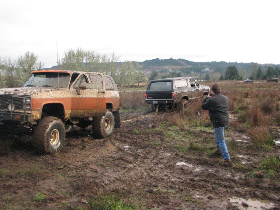 Mud-covered orange and white 1978 Ford Bronco with large tires, and a black 1978 Ford Bronco being pulled by a rope in a muddy field