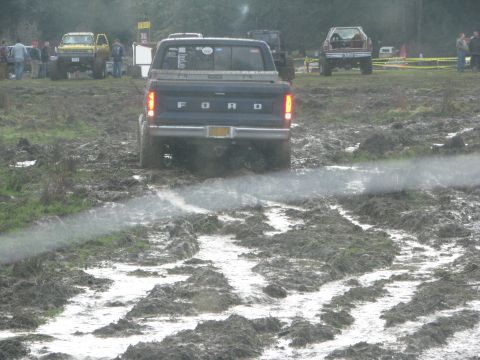 1978 Ford Bronco - A Ford pickup truck navigates a muddy track during an off-road event. Other vehicles and spectators are visible in the background.