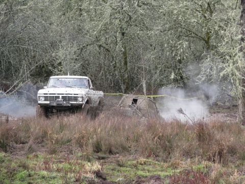 1978 Ford Bronco - A white Ford pickup truck navigates a muddy off-road course, with a small structure and yellow caution tape visible.