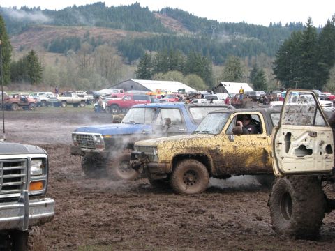 1978 Ford Bronco - Several off-road vehicles are engaged in a mud bogging event with spectators and buildings in the background.