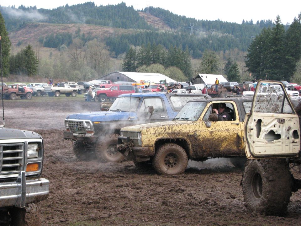 Blue Ford truck and yellow Chevy truck in mud pit, other vehicles parked in background