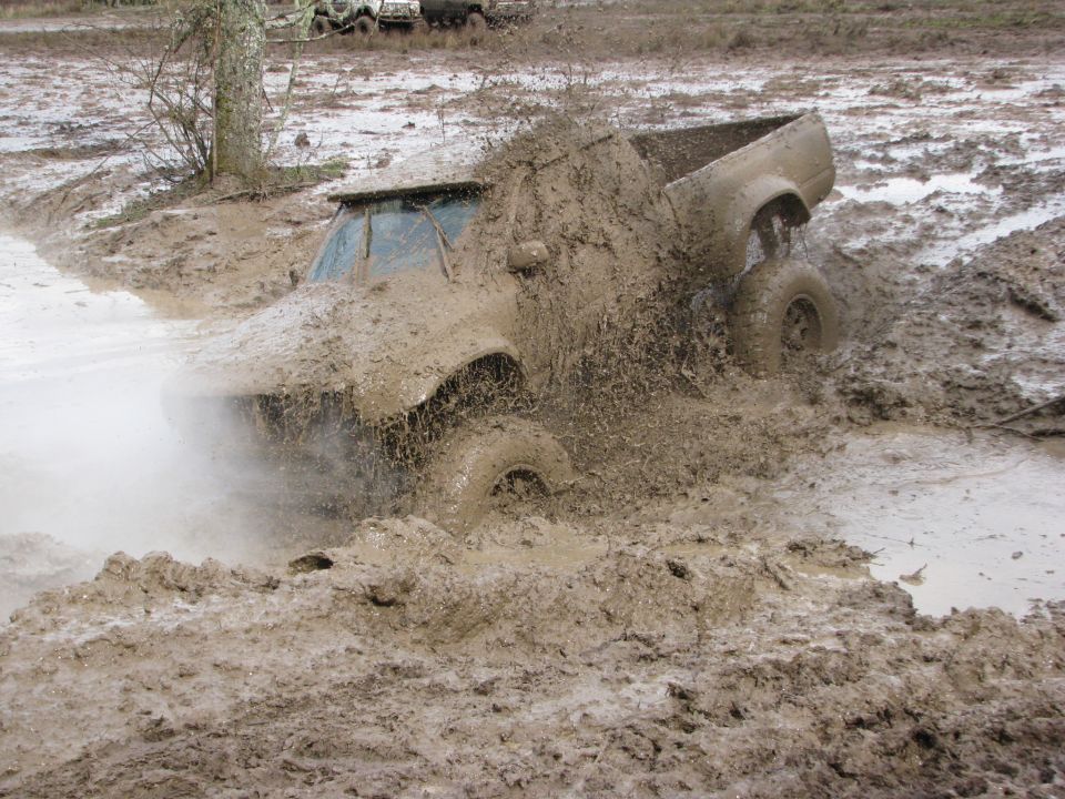 Off-road truck deep in mud, front end obscured by spray. Another vehicle visible in background