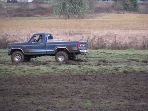 1978 Ford Bronco - A blue pickup truck navigates through a muddy, grassy area with a wooded background.