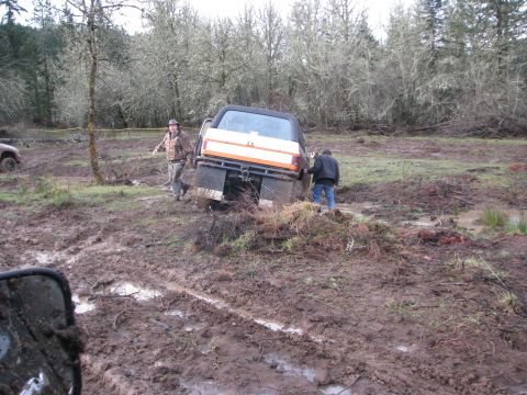 1978 Ford Bronco - Two people attempt to free an orange and white pickup truck that is stuck deep in the mud.