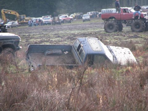 1978 Ford Bronco - A blue pickup truck is partially submerged in mud and tall grass, with other vehicles and an excavator visible in the background.