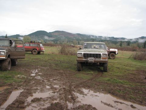 1978 Ford Bronco - Several off-road vehicles are parked in a muddy field under an overcast sky with hills in the distance.